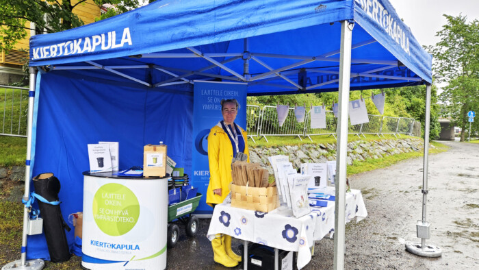 Kiertokapulan tapahtumateltassa seisoo nainen keltaisissa sadevarusteissa. Teltassa on esitteitä ja muita tiedotusmateriaaleja Kiertokapulan toiminnasta.